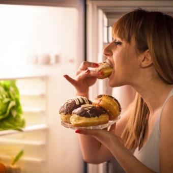 Una mujer come donuts con la nevera abierta