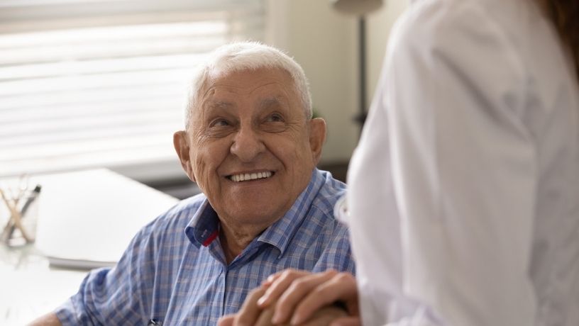 Hombre de tercera edad sonríe al tomar la mano de su cuidadora